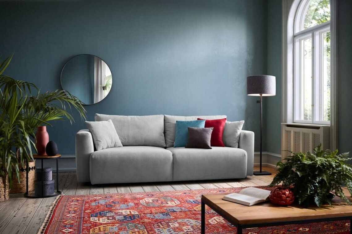 Graues Funktionssofa in einem stilvoll eingerichteten Wohnzimmer, frontal betrachtet, mit bunten Kissen und einem roten Teppich.