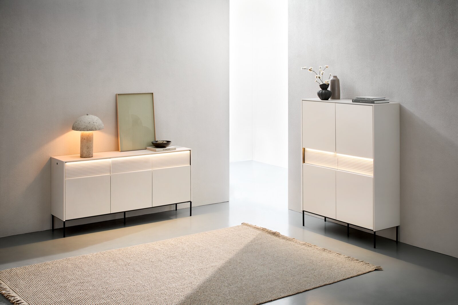 Minimalistisches Wohnambiente mit zwei hellbeigen Sideboards/Kommoden auf schwarzen Metallfüßen und integrierter LED-Lichtleiste, dekoriert mit Lampe, Bild, Vasen und Büchern Perspektive: frontale Ansicht in leichter Schrägansicht von links.