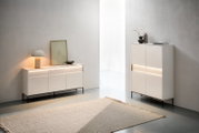 Minimalistisches Wohnambiente mit zwei hellbeigen Sideboards/Kommoden auf schwarzen Metallfüßen und integrierter LED-Lichtleiste, dekoriert mit Lampe, Bild, Vasen und Büchern; Perspektive: frontale Ansicht in leichter Schrägansicht von links.