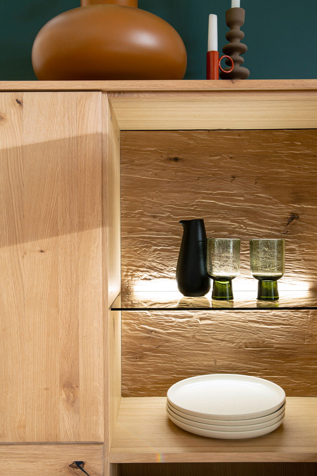 Detailansicht eines Holz-Sideboards mit dekorativen Elementen wie einer Vase, Kerzen und Gläsern auf einem beleuchteten Regalbrett.