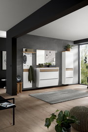 Modernes Flur-Set mit weißen Hängeschränken, Spiegel, Garderobenpaneel und Holz-Akzenten vor grauer Wand, fotografiert in leichter Schrägansicht von vorn.