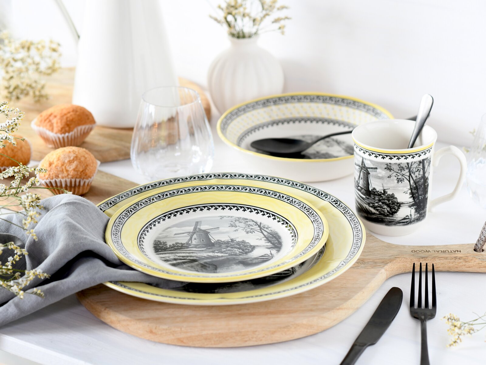 Gedeckter Frühstückstisch mit gelb-grau gemustertem Geschirrset (Teller, Schüssel und Tasse mit Windmühlenmotiv), Glas und Besteck auf Holzbrett, fotografiert in leicht erhöhter Frontansicht.
