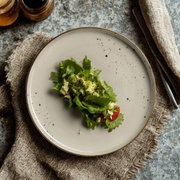 Grauer, gesprenkelter Keramikteller mit kleinem grünen Blattsalat und Tomatenstück, auf Leinentuch neben Besteck und Gläsern, fotografiert aus der Draufsicht (Top-Down).