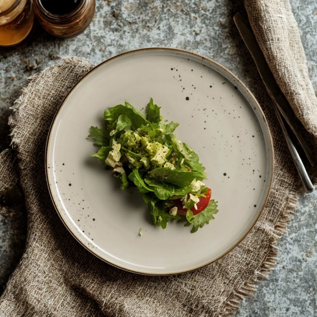 Grauer, gesprenkelter Keramikteller mit kleinem grünen Blattsalat und Tomatenstück, auf Leinentuch neben Besteck und Gläsern, fotografiert aus der Draufsicht (Top-Down).