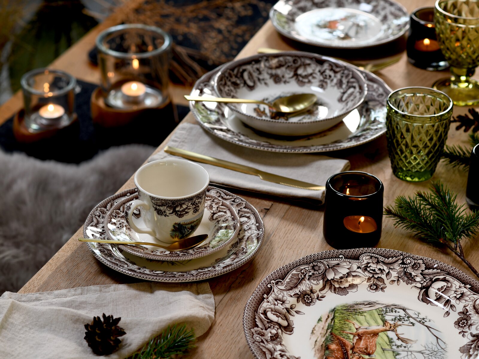 Festlich gedeckter Holztisch mit braun-weißem Porzellan-Geschirr (Teller, Schale und Tasse) mit Waldtier-Motiv, goldfarbenem Besteck, grünen Gläsern und Kerzenlicht, aufgenommen aus leicht schräger Draufsicht.