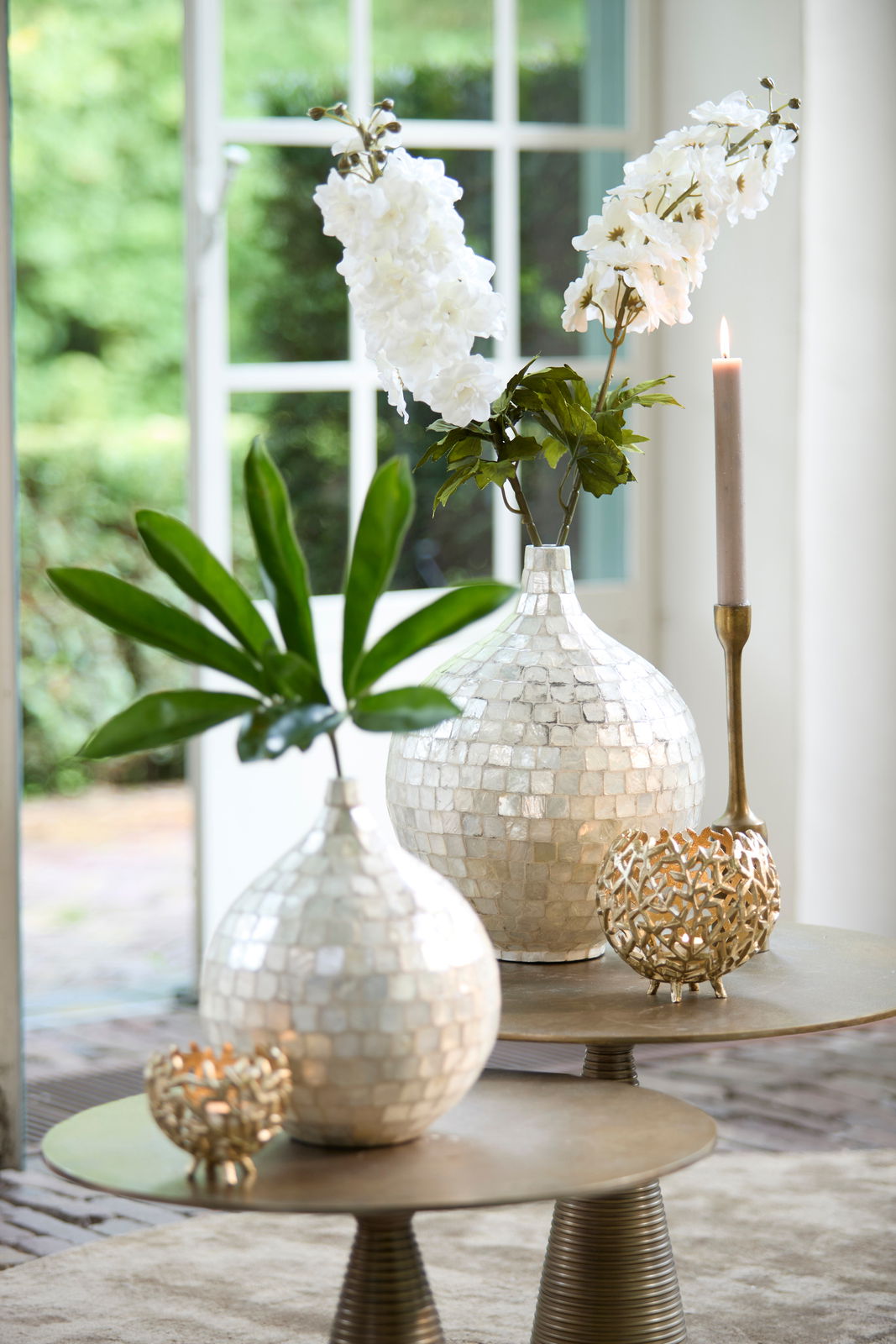 Elegantes Teelicht in goldener, durchbrochener Kugelform auf einem Tisch, umgeben von dekorativen Vasen und Kerzen, aus der Seitenperspektive fotografiert.