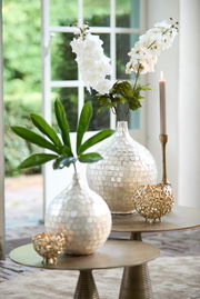 Elegantes Teelicht in goldener, durchbrochener Kugelform auf einem Tisch, umgeben von dekorativen Vasen und Kerzen, aus der Seitenperspektive fotografiert.