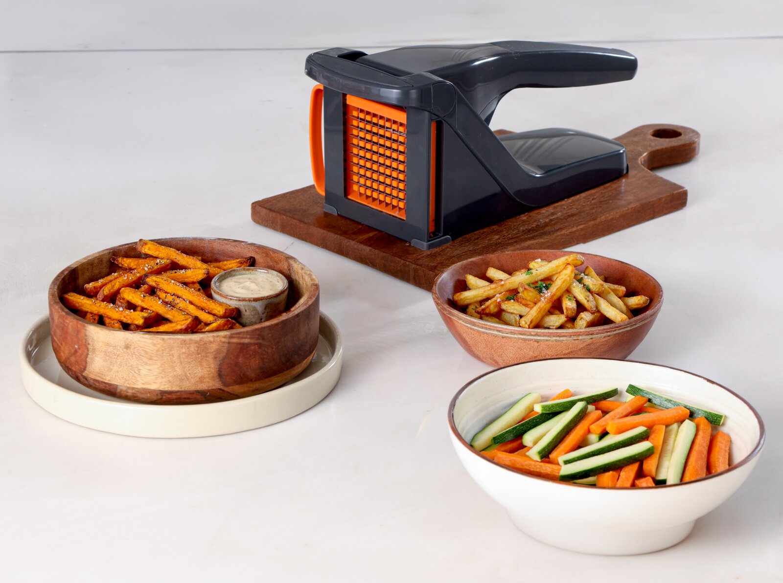 Schwarzer Pommes- und Gemüseschneider mit orangefarbenem Schneidegitter auf Holzbrett, schräg von vorn links fotografiert, daneben Schalen mit Pommes und Gemüsesticks