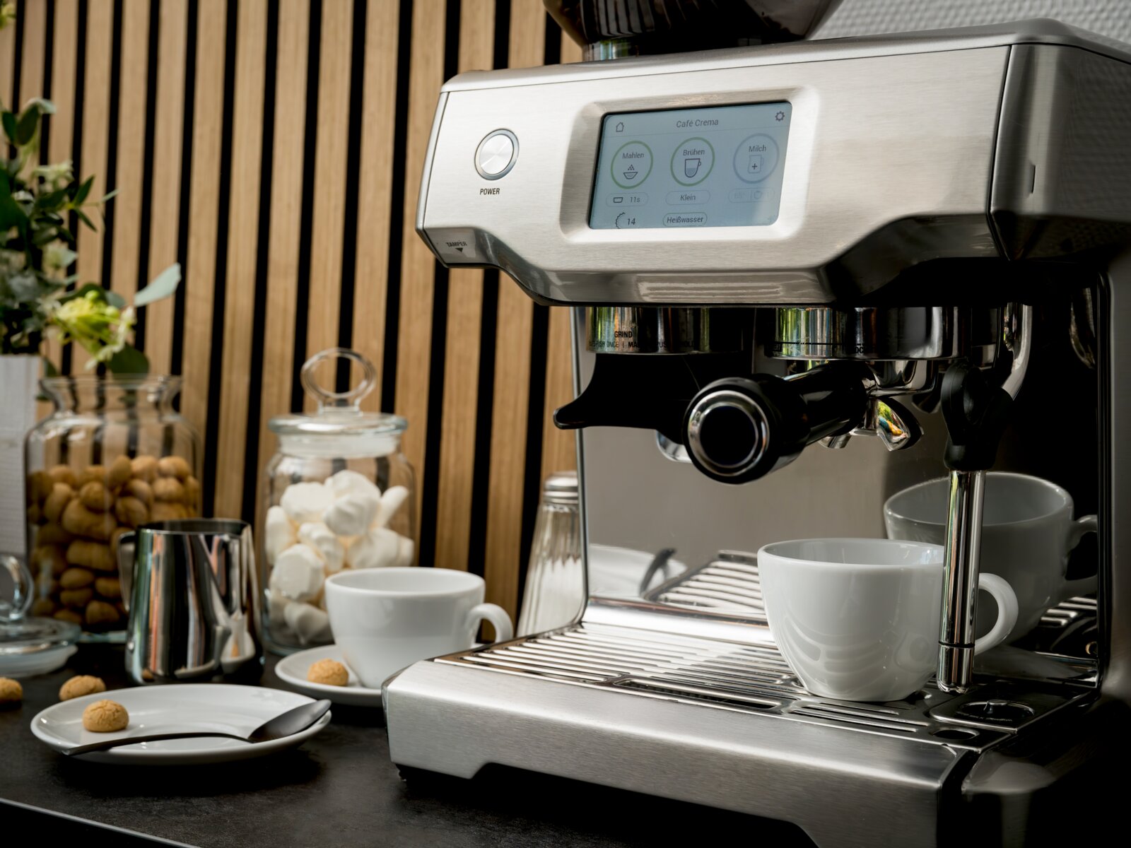 Edelstahl-Siebträgermaschine mit Touchdisplay und Dampflanze, frontal leicht von links auf der Küchenarbeitsplatte fotografiert; davor stehen weiße Tassen sowie Gläser mit Keksen und Marshmallows.