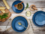 Dunkelblaues Keramikgeschirr-Set mit strukturierter Glasur auf rustikalem Holztisch, mit goldenem Besteck, Oliven, Brot und Kräutern, Draufsicht von oben