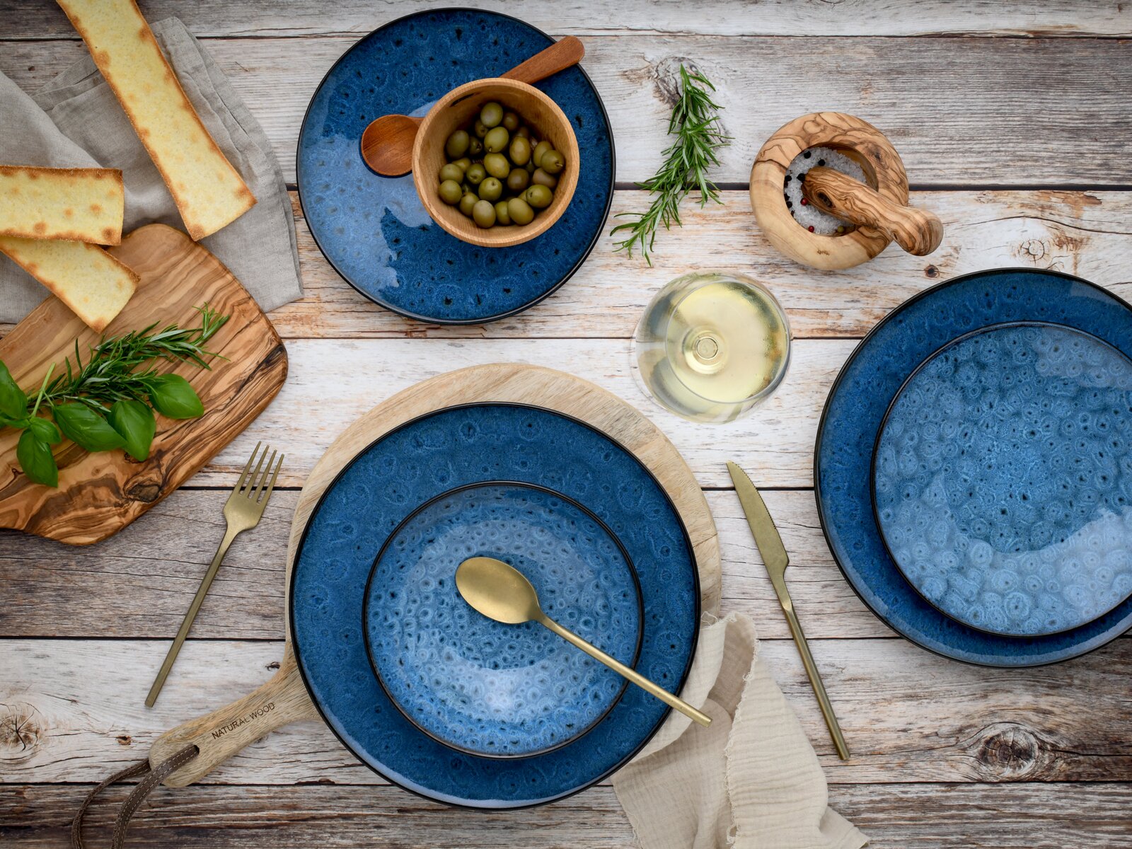 Dunkelblaues Keramikgeschirr-Set mit strukturierter Glasur auf rustikalem Holztisch, mit goldenem Besteck, Oliven, Brot und Kräutern, Draufsicht von oben