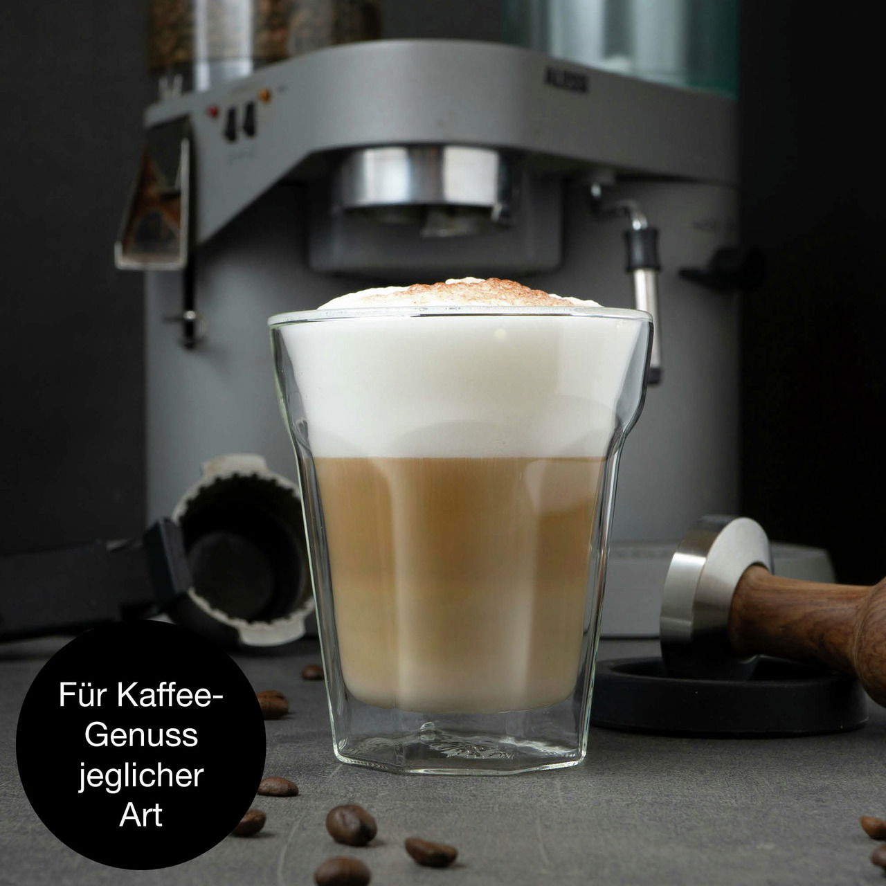 Doppelwandiges Glas mit Latte Macchiato, im Hintergrund eine Kaffeemaschine, seitliche Perspektive