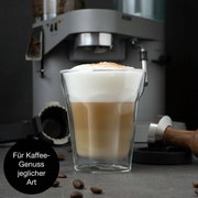 Doppelwandiges Glas mit Latte Macchiato, im Hintergrund eine Kaffeemaschine, seitliche Perspektive