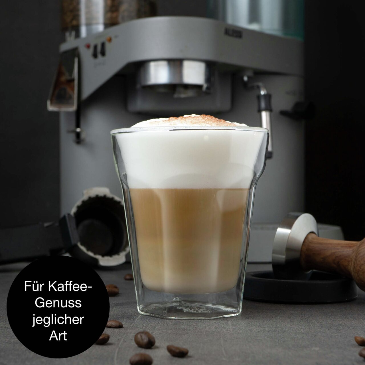 Doppelwandiges Glas mit Latte Macchiato, im Hintergrund eine Kaffeemaschine, seitliche Perspektive