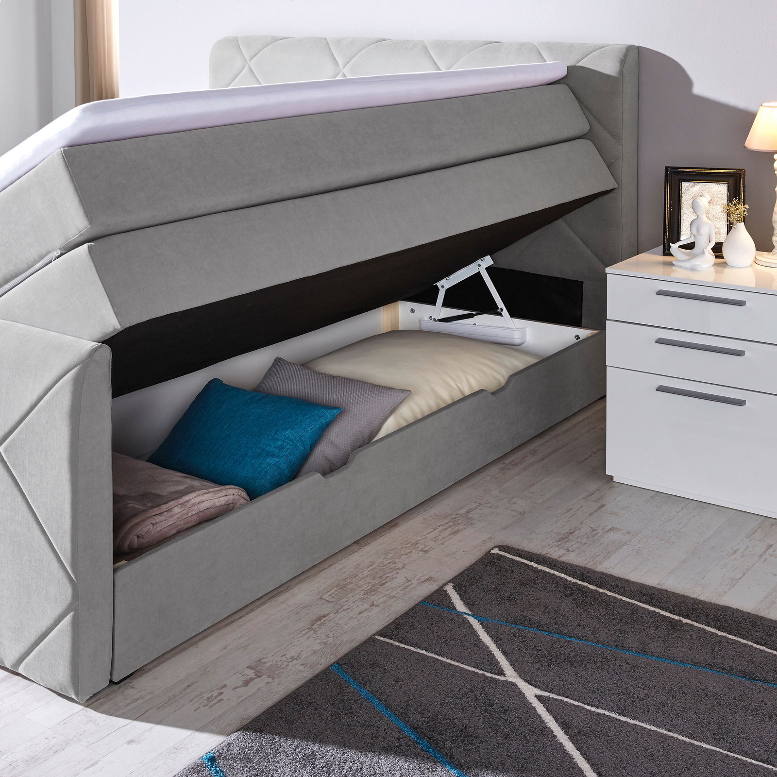 Boxspringbett mit geöffnetem Stauraum, seitliche Perspektive, graue Polsterung, Kissen und Decken im Inneren sichtbar.