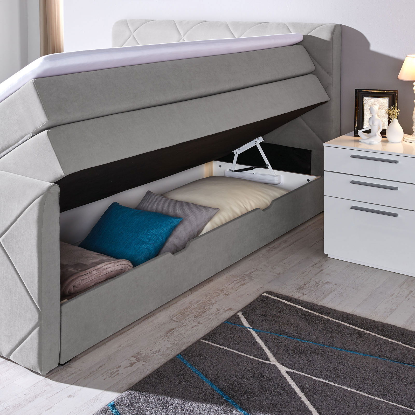 Boxspringbett mit geöffnetem Stauraum, seitliche Perspektive, graue Polsterung, Kissen und Decken im Inneren sichtbar.