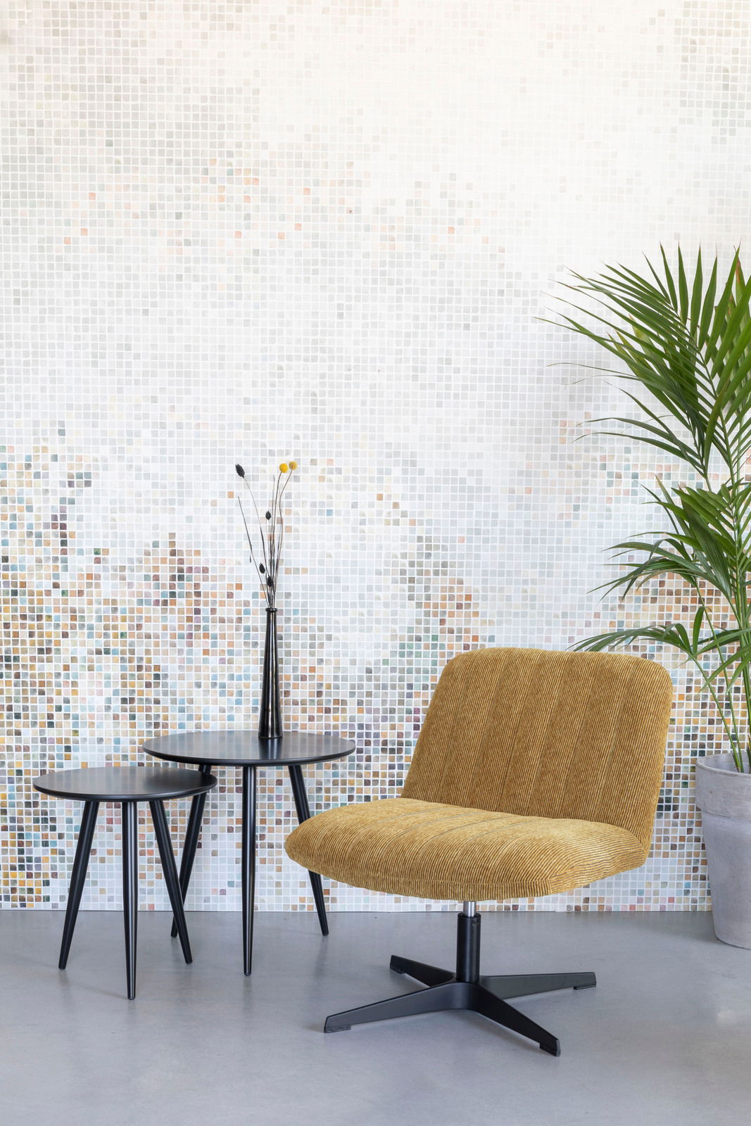 Moderner Sessel in Senfgelb mit schwarzem Metallfuß, seitliche Perspektive, vor einer Mosaikwand und neben zwei schwarzen Beistelltischen.