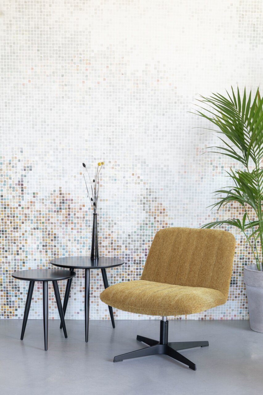 Z2 Sessel BELMONDO Moderner Sessel in Senfgelb mit schwarzem Metallfuß, seitliche Perspektive, vor einer Mosaikwand und neben zwei schwarzen Beistelltischen.