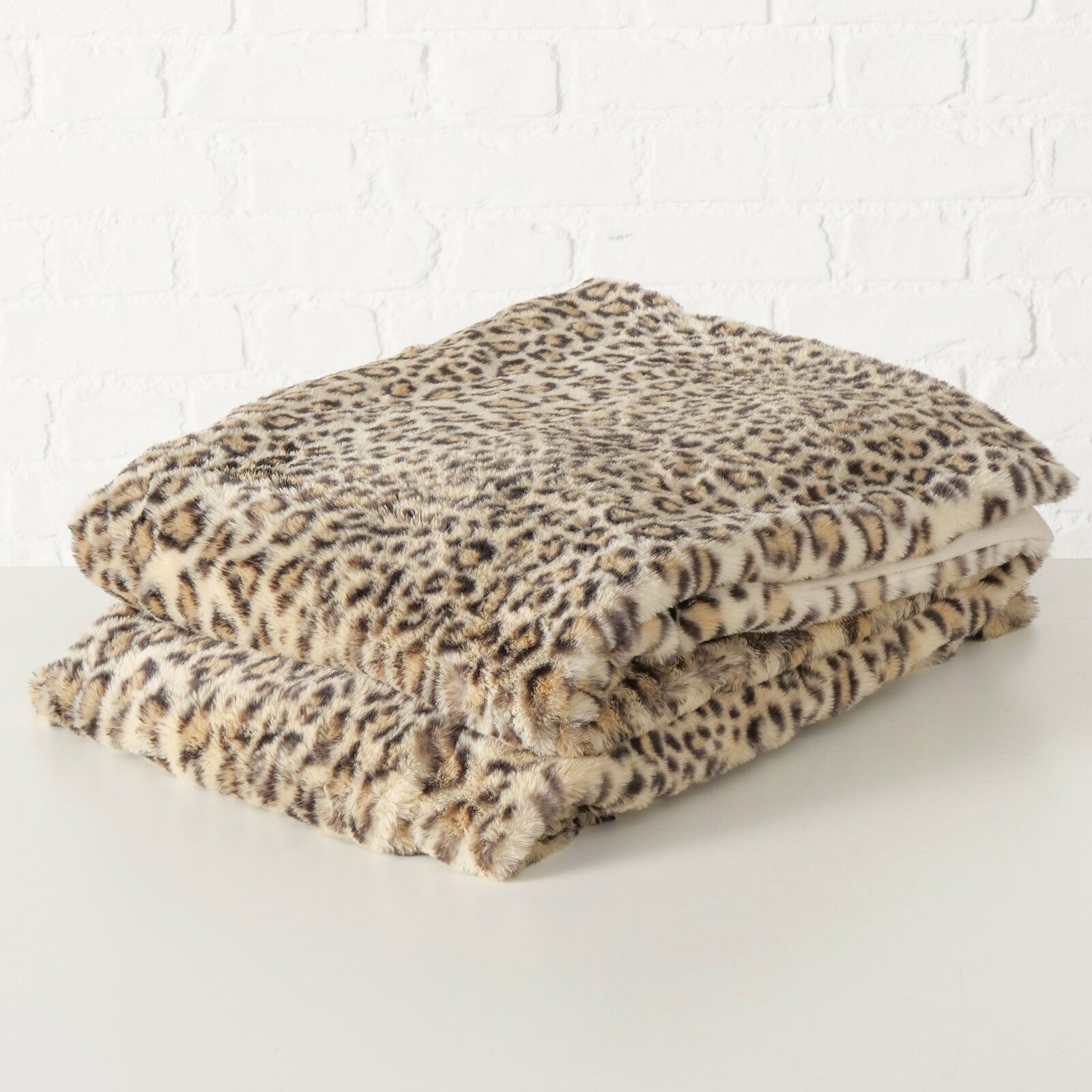 Leopardengemusterte Decke Leony aus weichem Material, gefaltet auf einem Tisch vor einer weißen Ziegelwand, seitliche Perspektive.