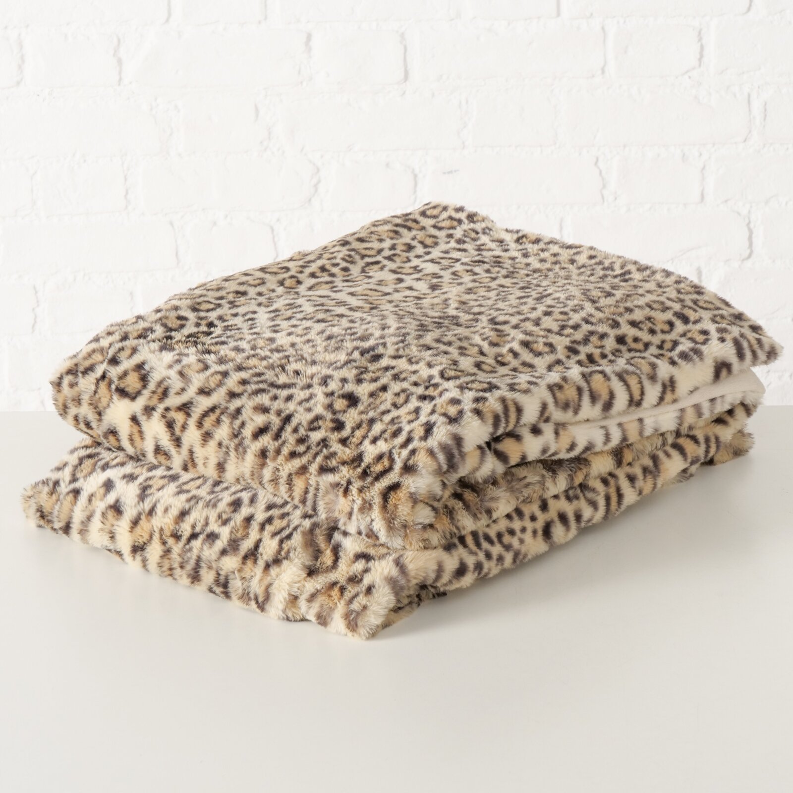 De Leony-deken met luipaardprint, gemaakt van zacht materiaal, opgevouwen op een tafel voor een witte bakstenen muur, gezien vanuit een zijdelingse hoek.