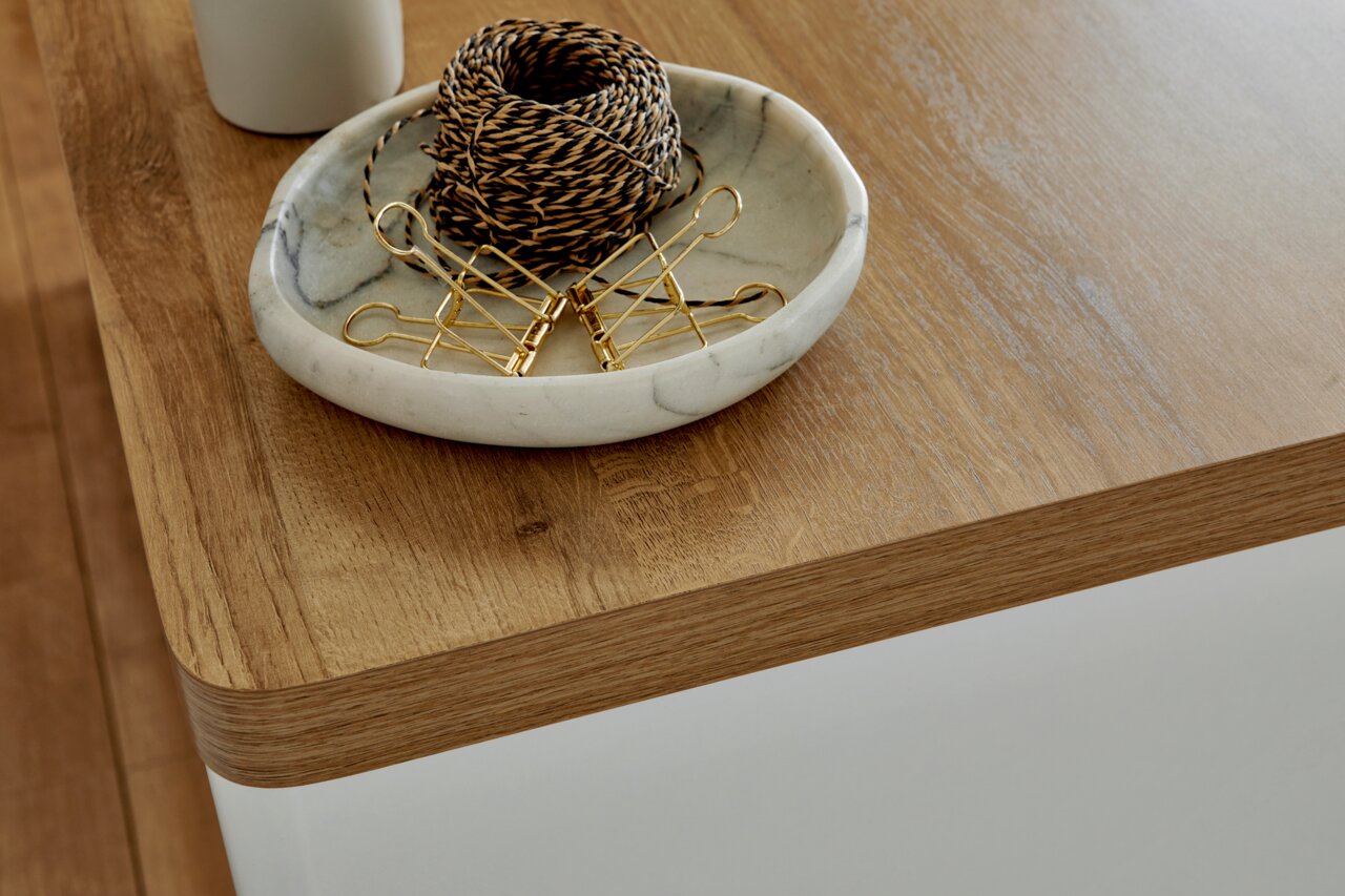 Nahaufnahme einer Ecke eines Eck-Schreibtisches aus Holz mit abgerundeten Kanten, darauf eine Schale mit Kordel und goldenen Klammern.