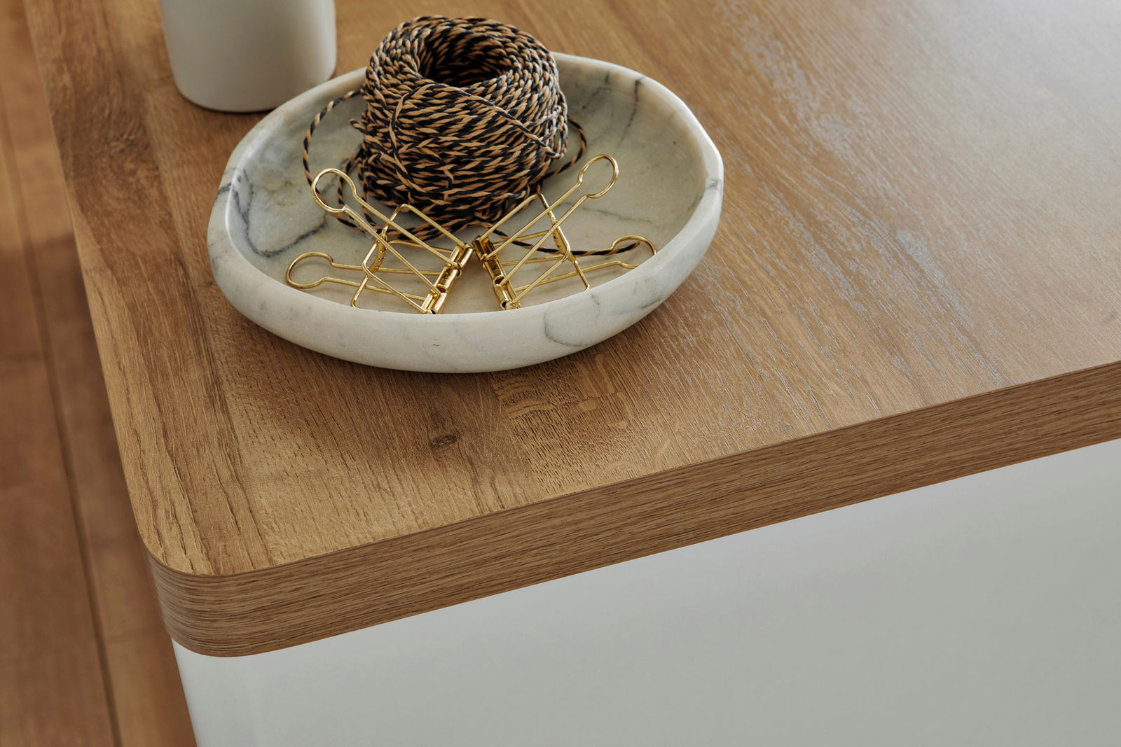 Nahaufnahme einer Ecke eines Eck-Schreibtisches aus Holz mit abgerundeten Kanten, darauf eine Schale mit Kordel und goldenen Klammern.