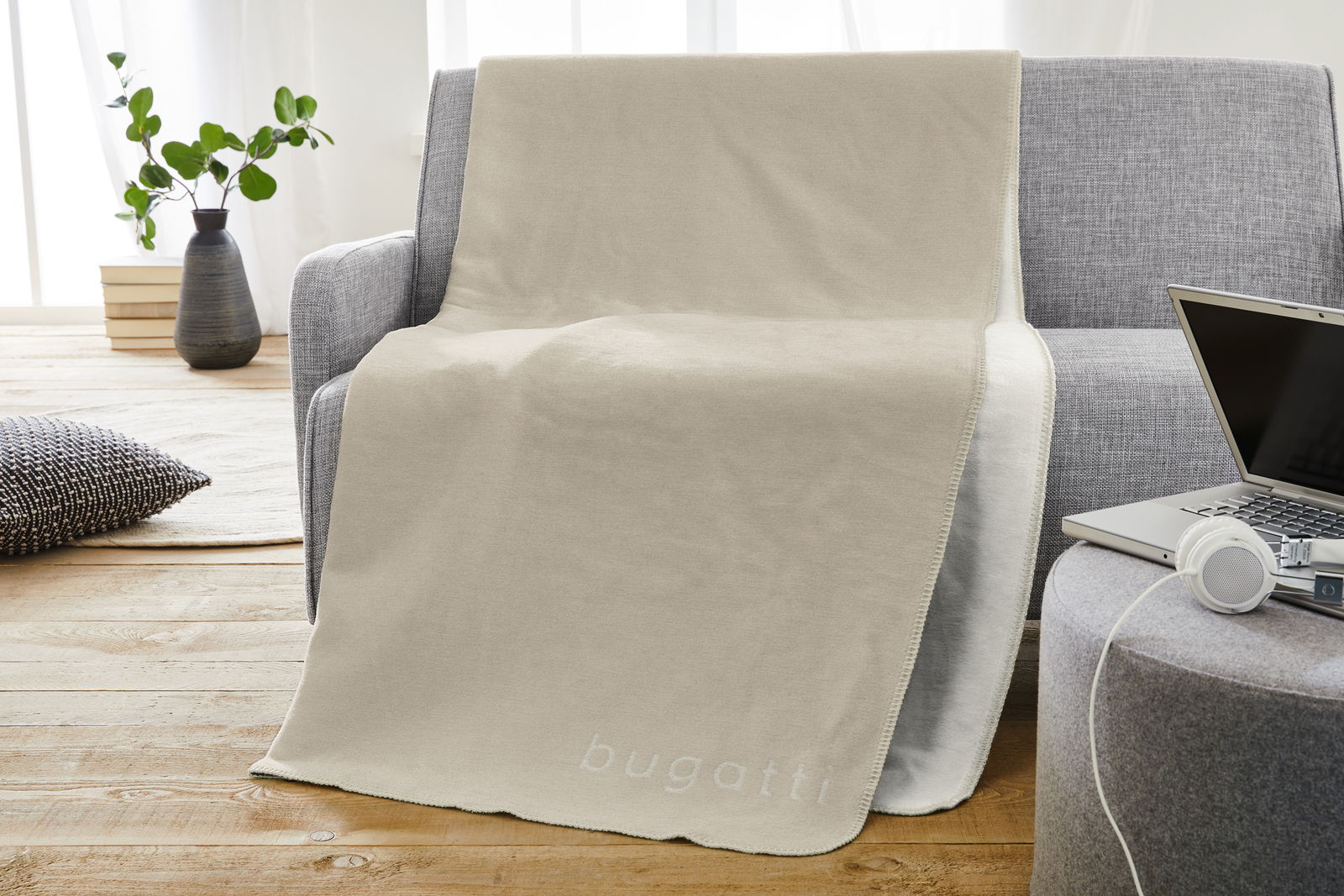 Beige Jacquard Decke von Bugatti auf einem grauen Sofa, mit einem modernen Wohnzimmer im Hintergrund. Perspektive von vorne.