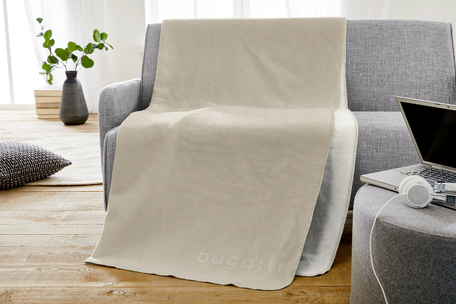 Bugatti Jacquard Decke Beige Jacquard Decke von Bugatti auf einem grauen Sofa, mit einem modernen Wohnzimmer im Hintergrund. Perspektive von vorne.