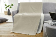 Beige Jacquard Decke von Bugatti auf einem grauen Sofa, mit einem modernen Wohnzimmer im Hintergrund. Perspektive von vorne.
