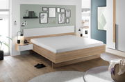 Modernes Schlafzimmer mit einem stilvollen Holzbett, flankiert von zwei Nachttischen mit Lampen. Die Wand ist mit Kunstwerken dekoriert. Perspektive: seitliche Ansicht des Bettes.