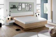 Moderne slaapkamer met een stijlvol houten bed, geflankeerd door twee nachtkastjes met lampjes. De muur is versierd met kunstwerken. Perspectief: zijaanzicht van het bed.
