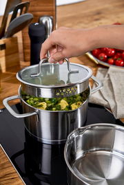 Hand hebt den Deckel eines Edelstahltopfs mit integriertem Abtropfsieb, gefüllt mit Gemüse, auf einem Herd. Perspektive von schräg oben.