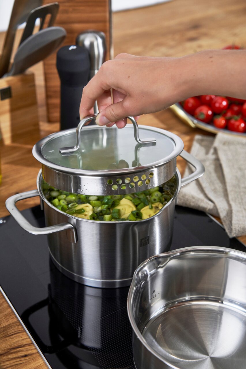 ZWILLING Topfset 5tlg. TRUE FLOW Hand hebt den Deckel eines Edelstahltopfs mit integriertem Abtropfsieb, gefüllt mit Gemüse, auf einem Herd. Perspektive von schräg oben.