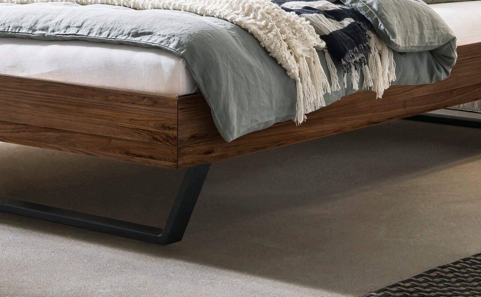 Modernes Bettgestell aus dunklem Holz mit schwarzem Metallfuß, mit grauer Bettwäsche und Decke; Detailansicht aus seitlicher, leicht schräger Perspektive
