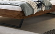 Modernes Bettgestell aus dunklem Holz mit schwarzem Metallfuß, mit grauer Bettwäsche und Decke; Detailansicht aus seitlicher, leicht schräger Perspektive