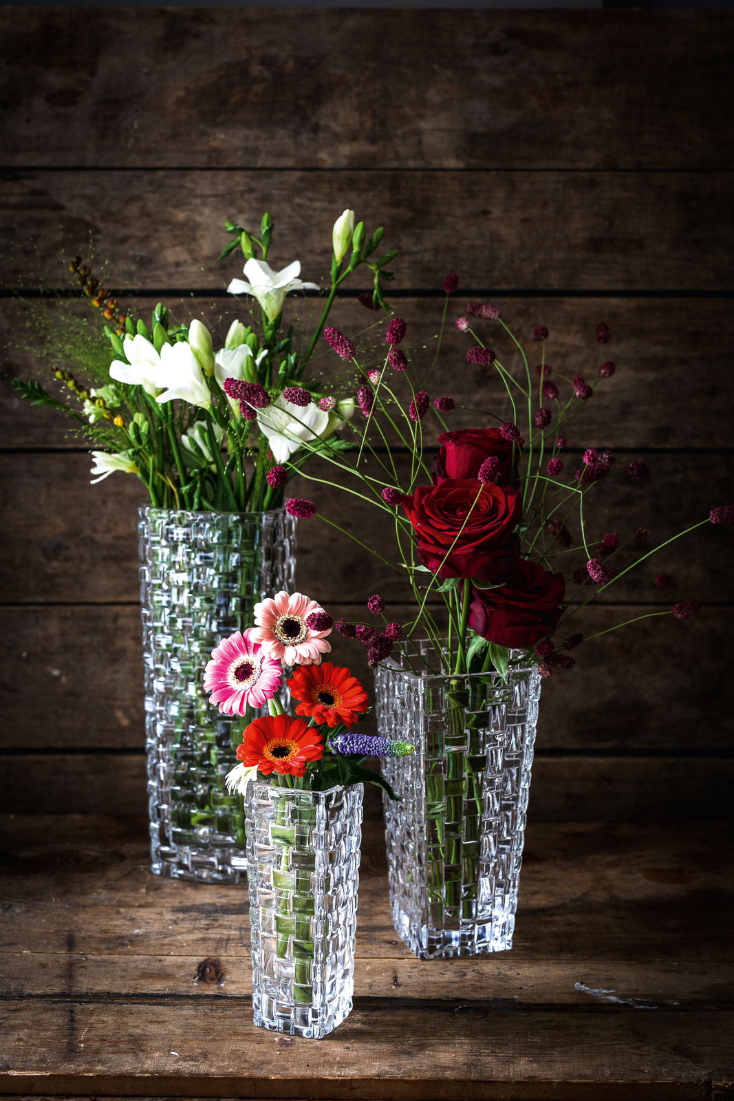 Drei elegante, klare Glasvasen mit Blumenarrangements, fotografiert aus einer leicht erhöhten Perspektive vor einem rustikalen Holzhintergrund.