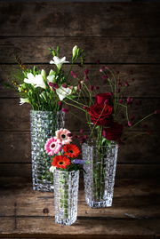 Drei elegante, klare Glasvasen mit Blumenarrangements, fotografiert aus einer leicht erhöhten Perspektive vor einem rustikalen Holzhintergrund.