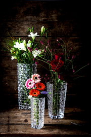 Drei elegante, klare Glasvasen mit Blumenarrangements, fotografiert aus einer leicht erhöhten Perspektive vor einem rustikalen Holzhintergrund.