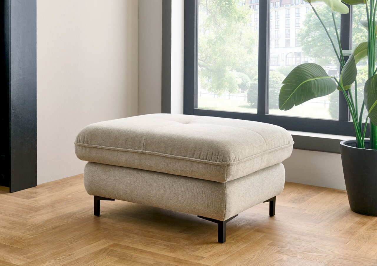 Beiger XL Hocker in einem modernen Raum, seitliche Perspektive, mit schwarzem Metallgestell und Holzfußboden.