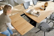 Aus schräger Vogelperspektive: moderner ausziehbarer Esstisch aus hellem Holz mit Ansteckplatte und schwarzem Gestell, umgeben von grauen Polsterstühlen; eine Person schiebt die Tischplatte.