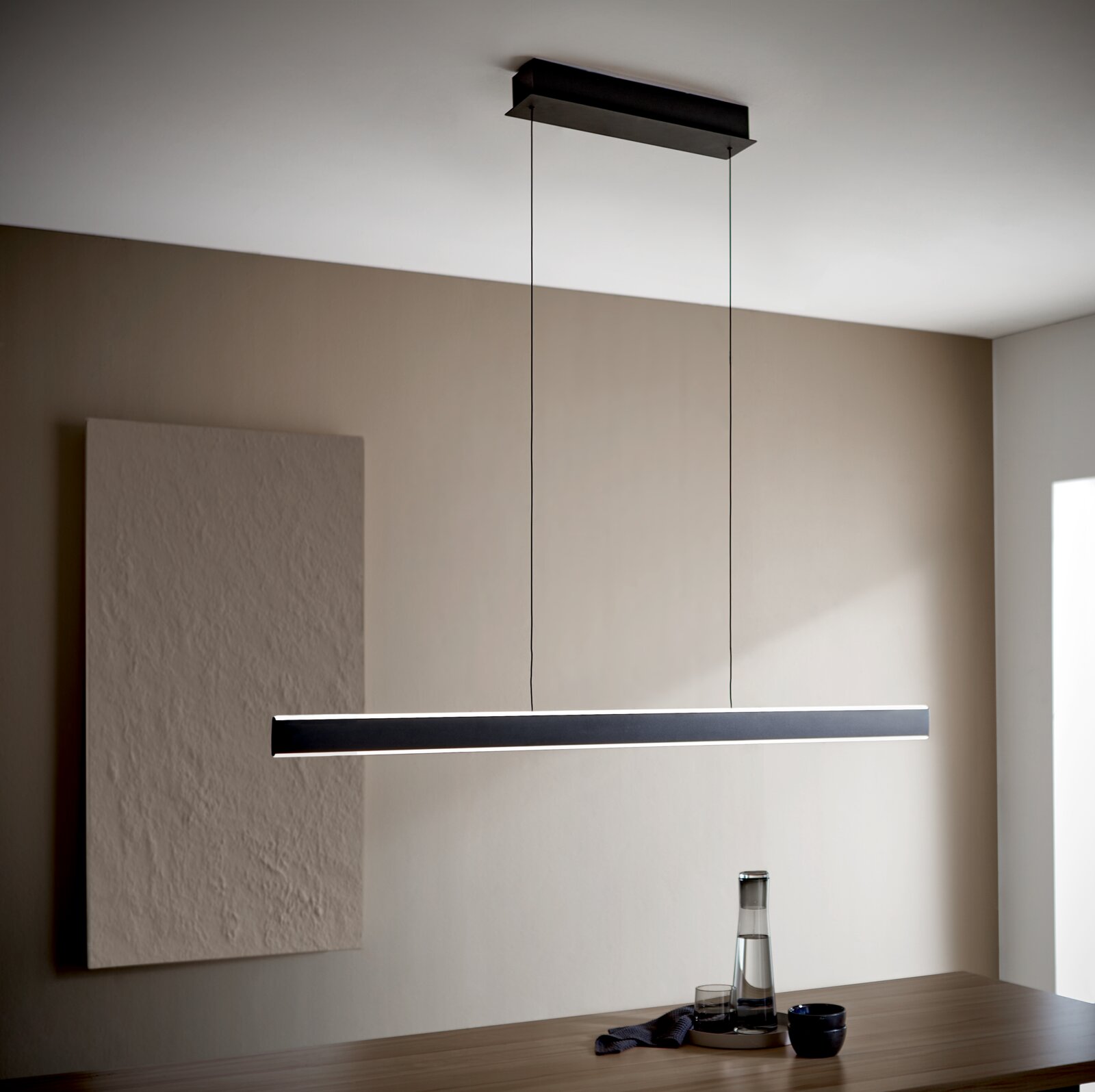 Moderne schwarze LED-Pendelleuchte in schlankem, länglichem Design, an zwei Kabeln von einer rechteckigen Deckenhalterung über einem Esstisch hängend perspektivische Ansicht leicht von unten.