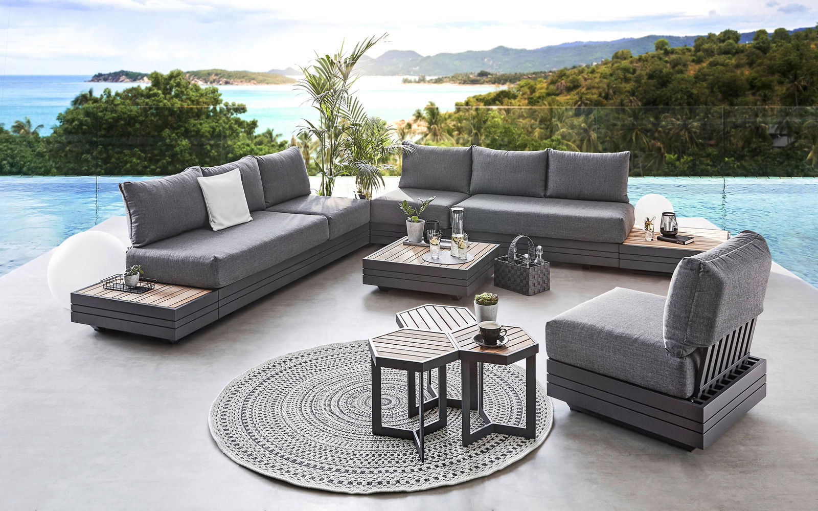 Moderne Outdoor-Lounge mit grauen Polstermöbeln und Holztischen, umgeben von tropischer Landschaft und Meerblick, aus der Vogelperspektive.