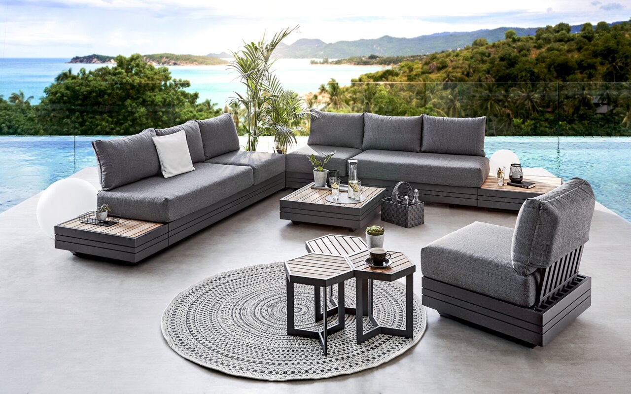 Moderne Outdoor-Lounge mit grauen Polstermöbeln und Holztischen, umgeben von tropischer Landschaft und Meerblick, aus der Vogelperspektive.