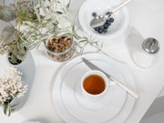 Helle Frühstückstafel in Draufsicht: weißes Geschirr mit Teetasse auf Teller, Schale mit Blaubeeren und Löffel, Glas mit Müsli sowie Zuckerdose und Blumen in Vase auf weißem Tisch.