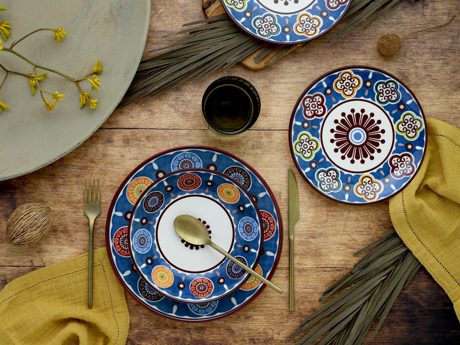Bunt gemustertes Keramikgeschirr-Set in Blau mit Teller und Schalen auf rustikalem Holztisch, dekoriert mit goldfarbenem Besteck und gelben Servietten, Draufsicht von oben.