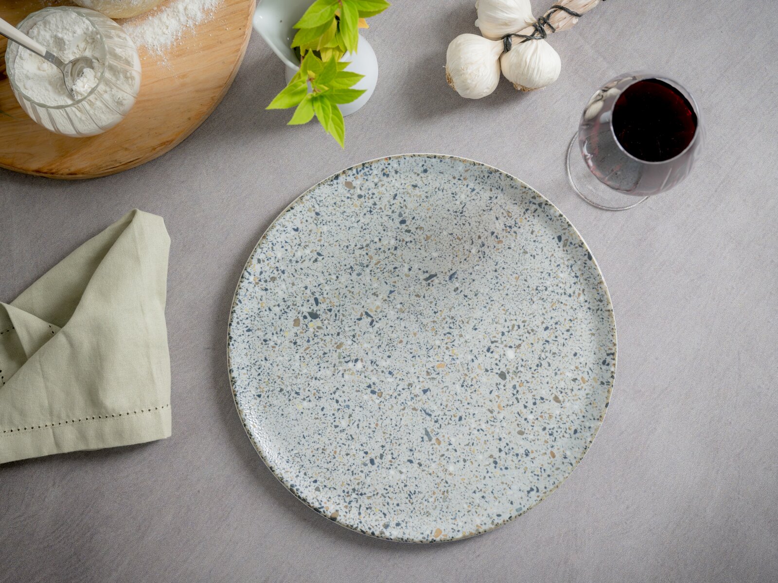 Runder, gesprenkelter Keramik-Speiseteller in Hellgrau/Beige auf grauer Tischdecke, Draufsicht (Top-Down), umgeben von Serviette, Basilikum, Knoblauch und einem Glas Rotwein