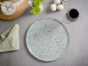 Runder, gesprenkelter Keramik-Speiseteller in Hellgrau/Beige auf grauer Tischdecke, Draufsicht (Top-Down), umgeben von Serviette, Basilikum, Knoblauch und einem Glas Rotwein