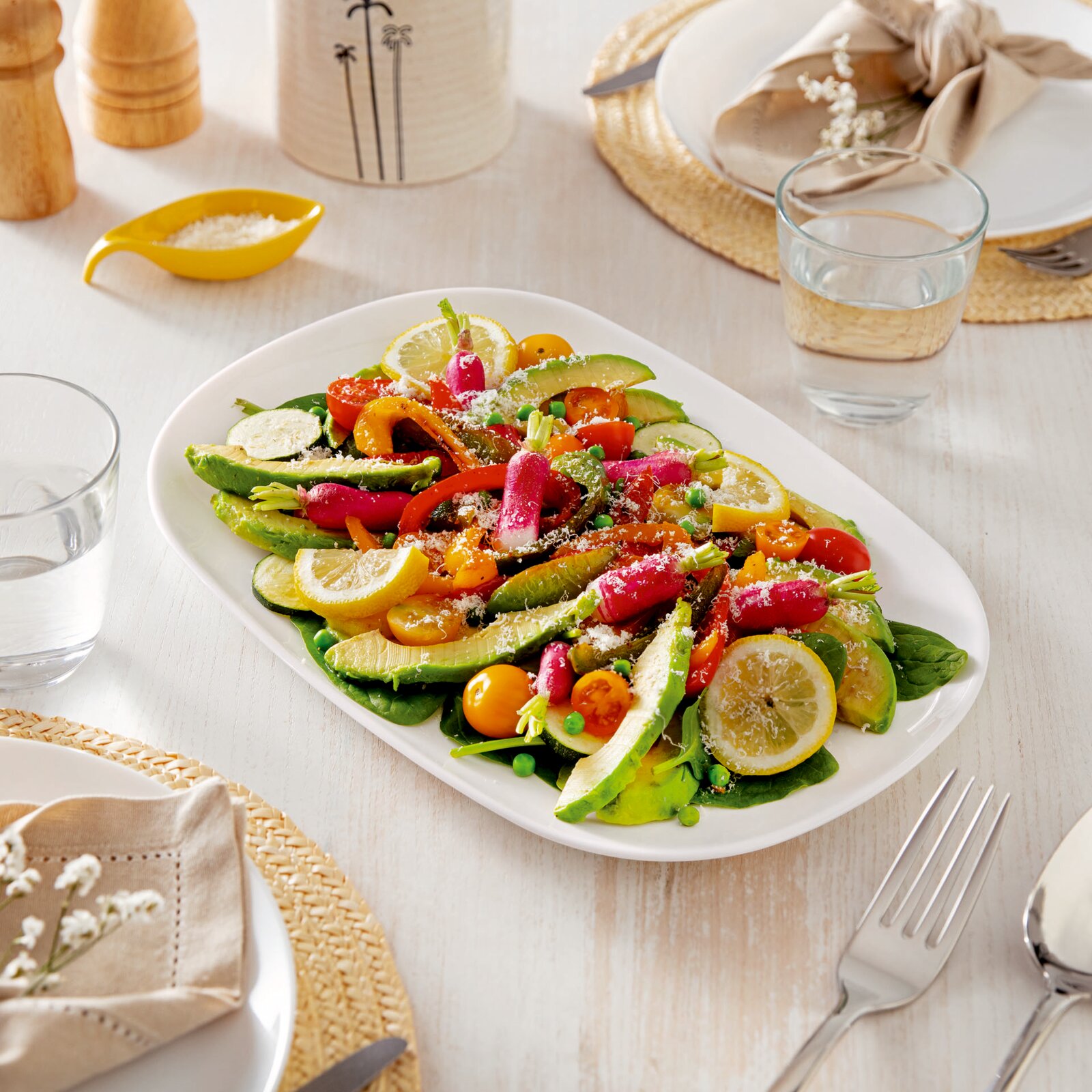 Bunte Gemüseplatte auf weißer Servierplatte mit Avocado, Radieschen, Kirschtomaten, Zucchini, Paprika, Erbsen und Zitronenscheiben, auf gedecktem Tisch – Perspektive: leicht von oben, schräg von vorn.