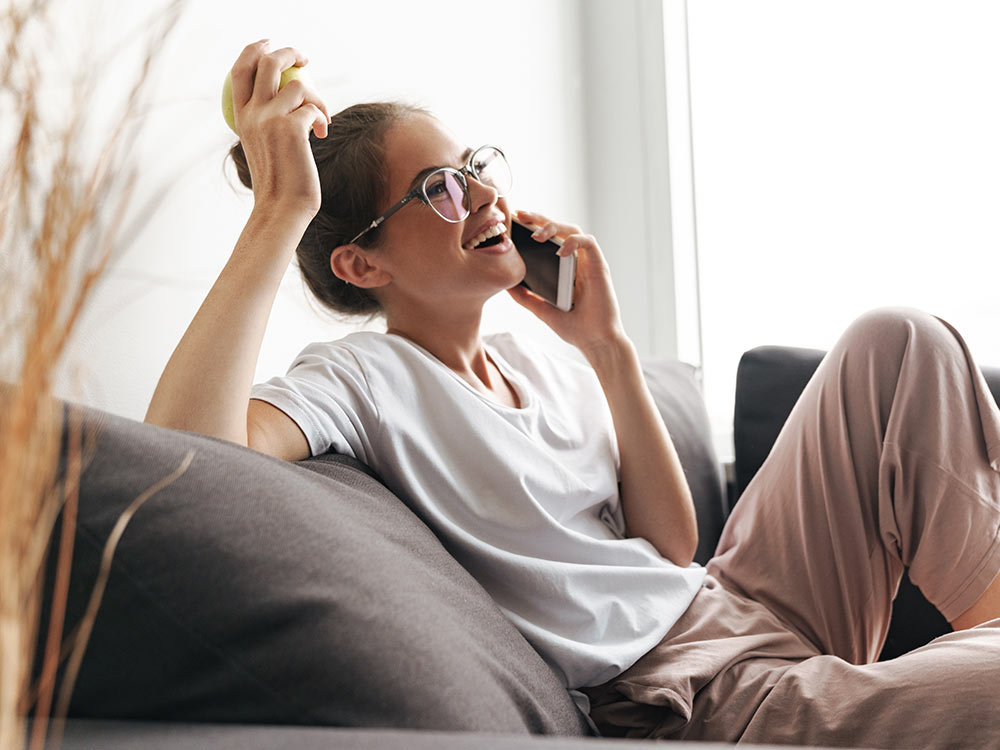 Een jonge vrouw met een bril en bruin haar zit ontspannen op een donkere bank, houdt een smartphone tegen haar oor en lacht. In haar andere hand houdt ze een appel vast. Ze draagt een wit T-shirt en een beige broek. Op de achtergrond is een licht raam te 