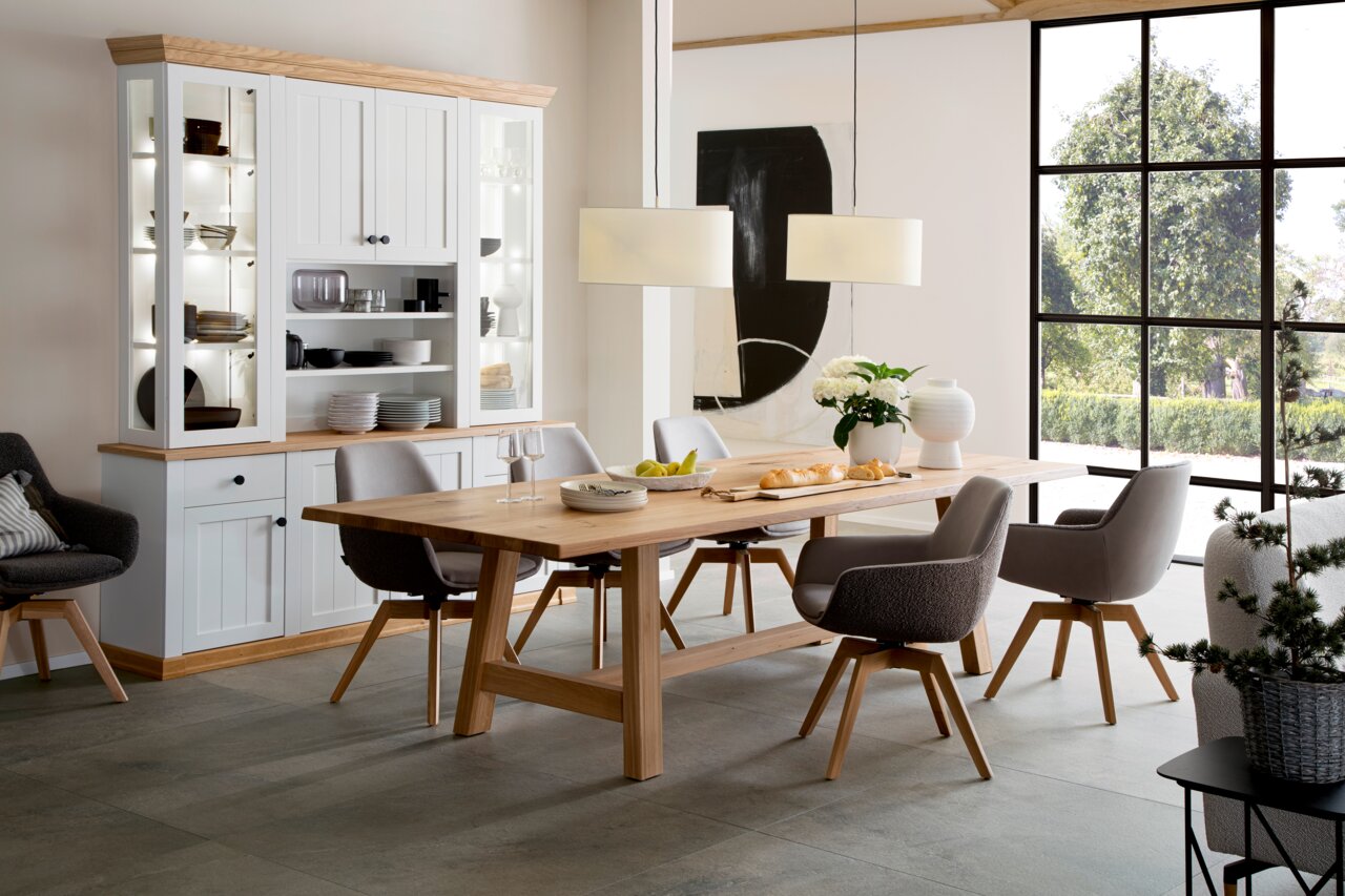 Moderne eethoek met een grote houten tafel en grijze stoelen, op de achtergrond een witte vitrinekast en grote ramen met uitzicht op het groen, zijaanzicht.
